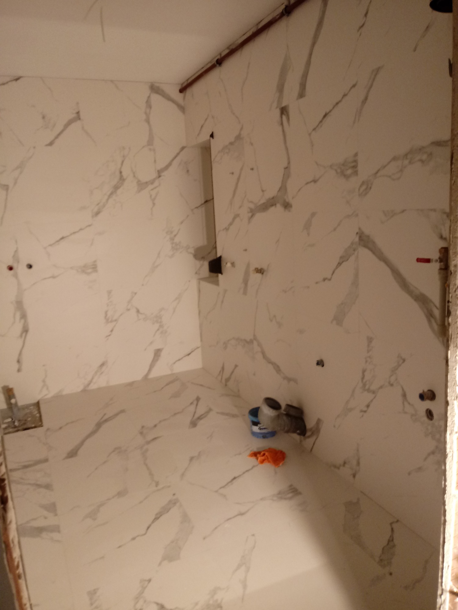 Wnętrze łazienki w trakcie prac wykończeniowych, widoczne płytki imitujące marmur na ścianach i podłodze, niezamontowana armatura i narzędzia glazurnicze.