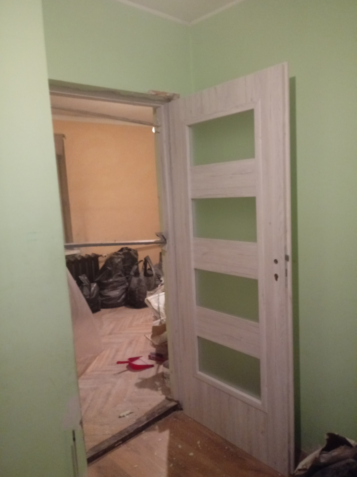 Otwarta, biała, drewnopodobna ościeżnica z czterema przeszkleniami, widok na remontowane wnętrze z workami gruzu i narzędziami, ściany w odcieniach zieleni i beżu.