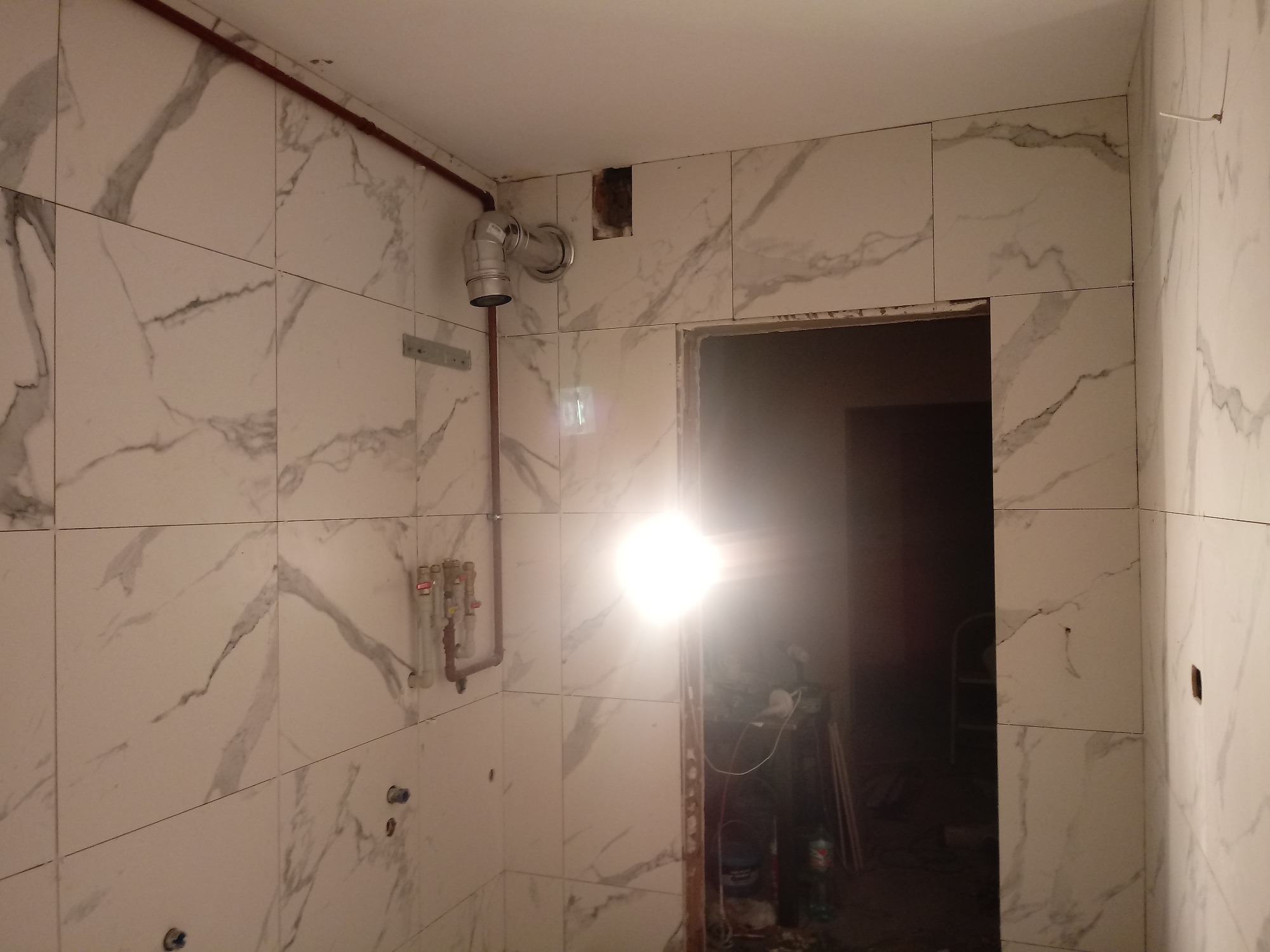 Ściany łazienki wyłożone białymi płytkami imitującymi marmur, widoczne rury instalacji wodnej i otwór w ścianie, przygotowane do montażu armatury.