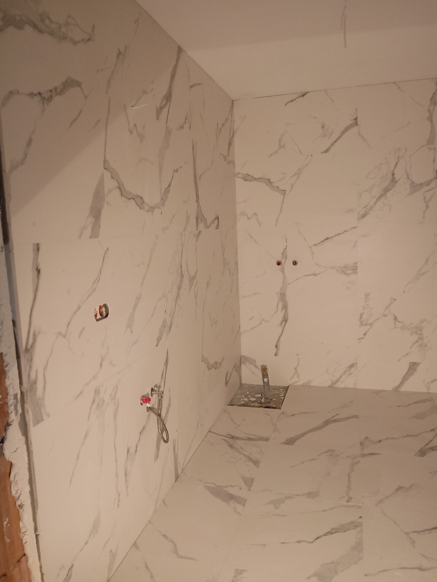 Białe płytki imitujące marmur na ścianach i podłodze w trakcie montażu, widoczne przyłącza wodne i otwory na gniazdka, ślady zaprawy na krawędzi ściany.