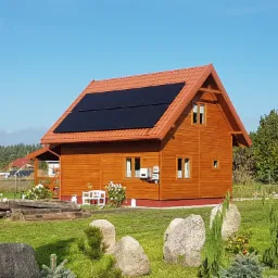 Drewniany dom z czerwoną dachówką i panelami słonecznymi na dachu, otoczony zielonym trawnikiem i dekoracyjnymi kamieniami.