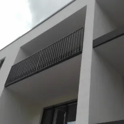 Nowoczesny budynek z białym tynkiem, balkonem z metalową balustradą i ciemnymi ramami okiennymi, widziany z dołu pod kątem.