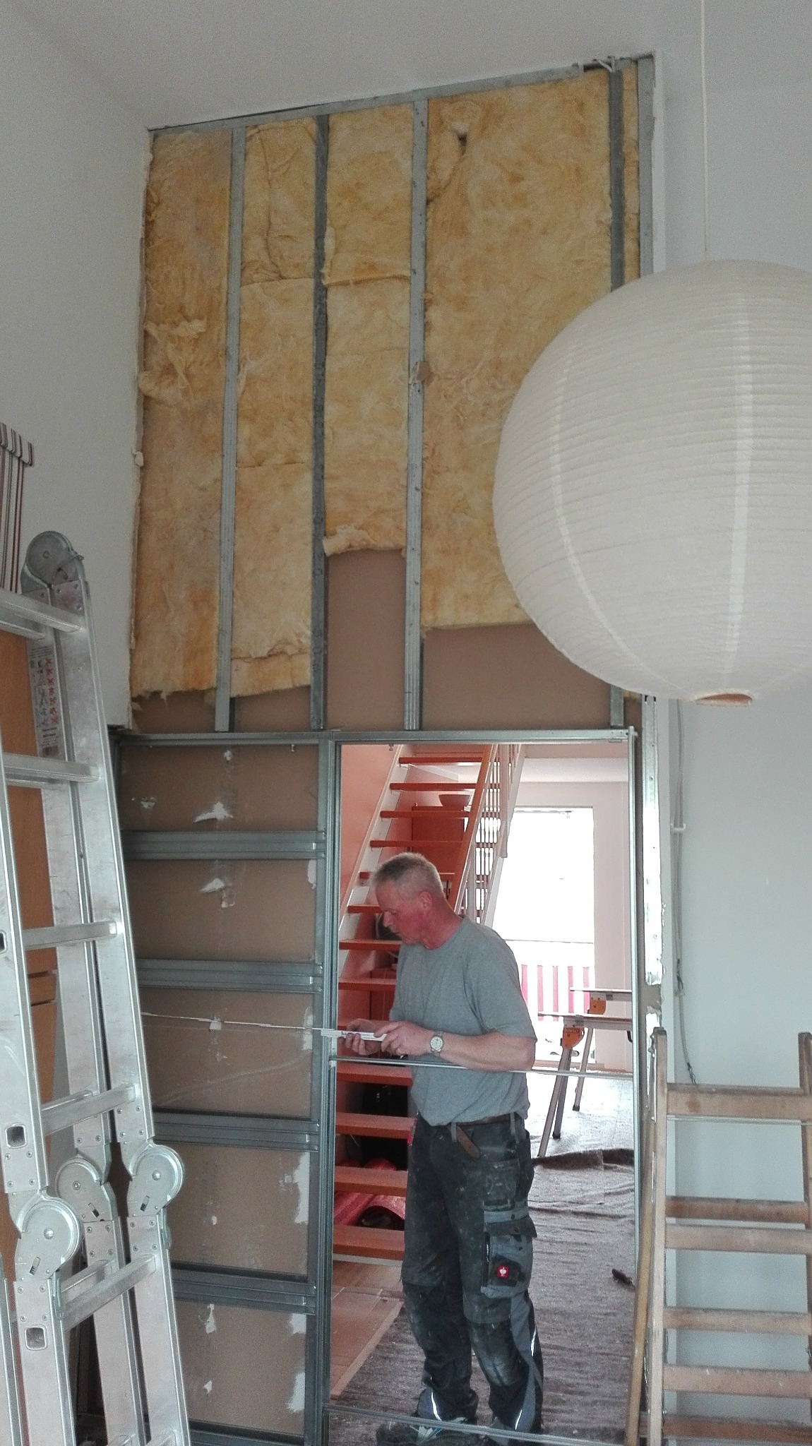 Pracownik mierzy metalową konstrukcję ściany działowej z izolacją z wełny mineralnej, widoczna drabina i schody w tle.