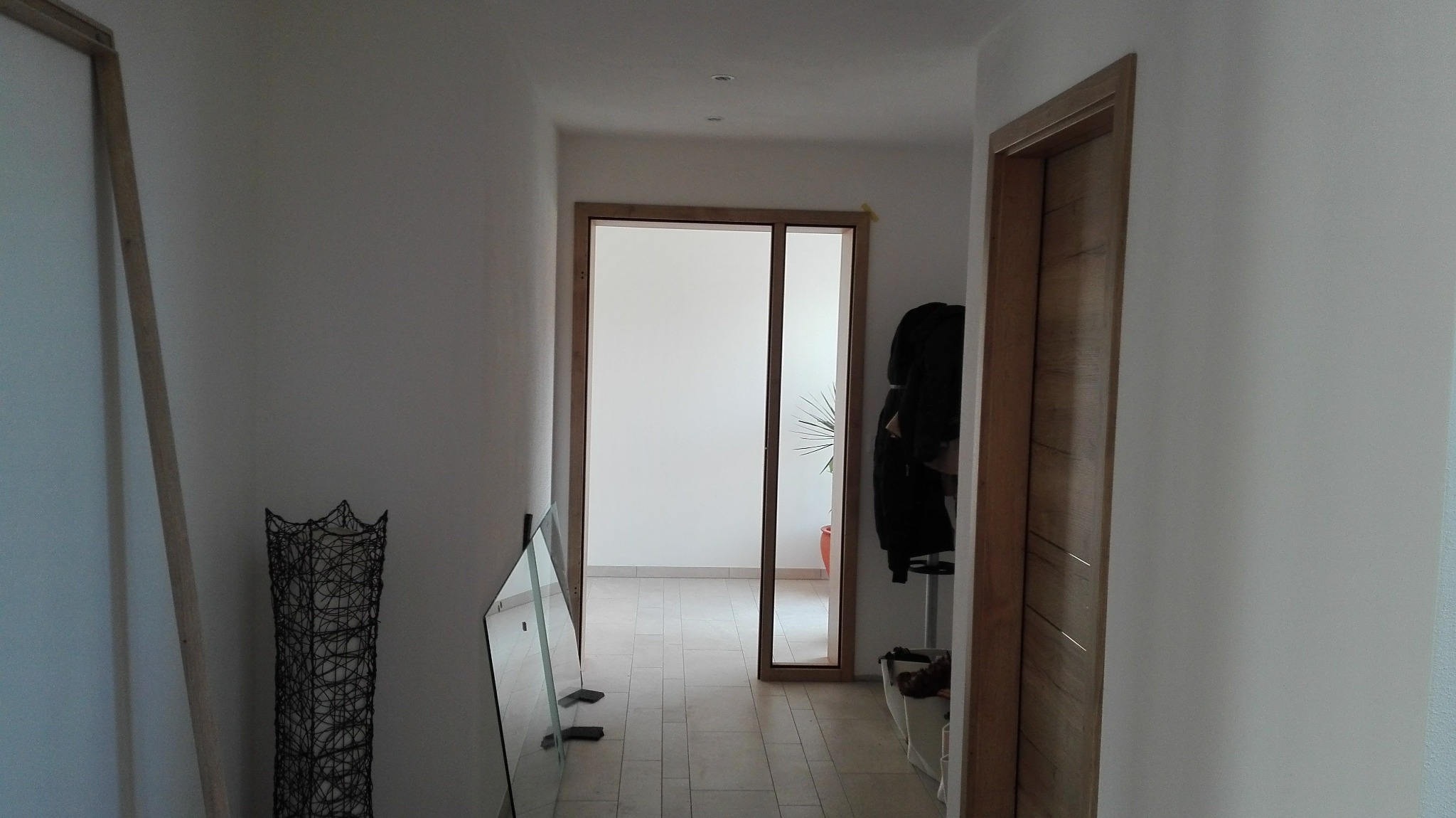 Korytarz w trakcie remontu: widoczne niezamontowane drzwi z drewnianą ramą, lustro oparte o ścianę, dekoracyjna lampa i fragment ściany po świeżym malowaniu.