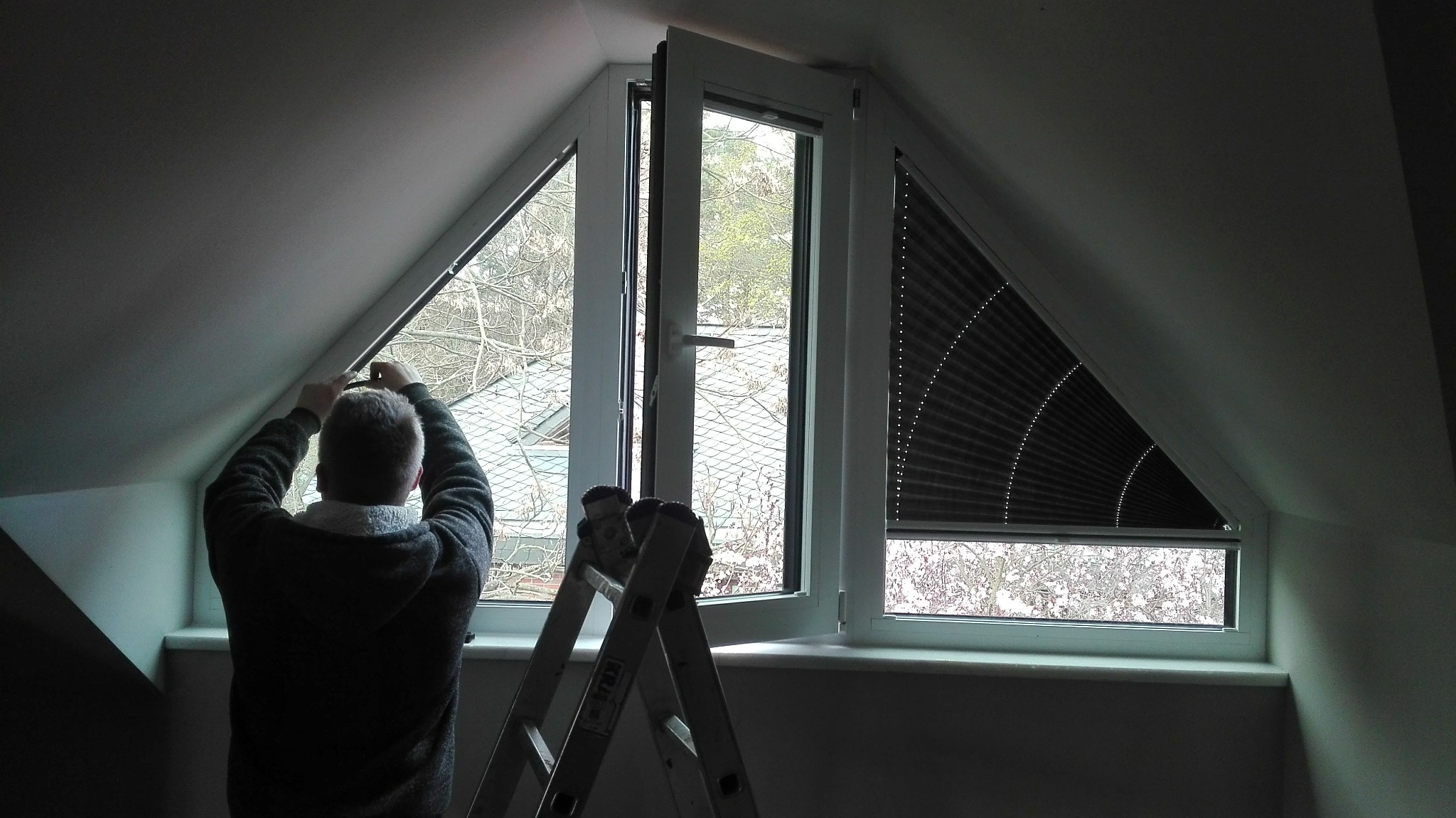 Pracownik montuje plisę okienną w trójkątnym oknie na poddaszu w Słupsku, widoczna drabina i uchylone okno.