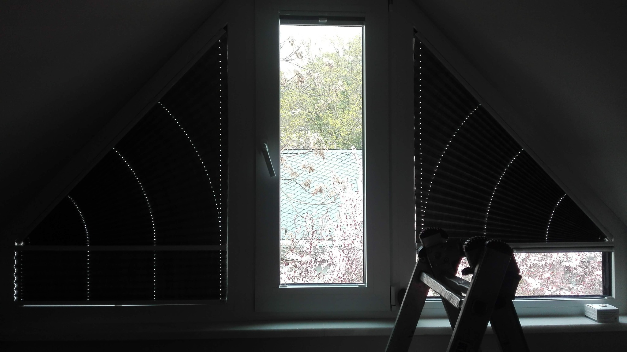 Okno strychowe z zamontowanymi roletami plisowanymi w kształcie trójkąta, drabina oparta o parapet, widok na drzewa i dach budynku za oknem.