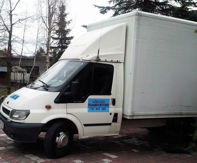 Biały samochód dostawczy z zabudową kontenerową, zaparkowany na brukowanej powierzchni. Na drzwiach widoczny niebieski napis z numerem telefonu. Tło stanowią drzewa i budynek.