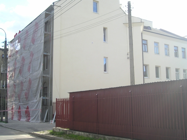 Budynek wielopiętrowy w trakcie renowacji elewacji, zabezpieczony siatką ochronną i rusztowaniami, widoczny fragment metalowego ogrodzenia.