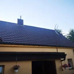 Dom z ciemnobrązowym dachem z blachy falistej, kominem i anteną satelitarną, widok z dołu na błękitne niebo.