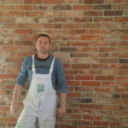 martom-lux - Mężczyzna w białym kombinezonie malarskim z logo firmy stoi na tle ściany z cegły klinkierowej w odcieniach czerwieni i brązu. Widoczne gniazdka elektryczne w ścianie.