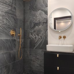martom-lux - Nowoczesna łazienka z szarymi płytkami imitującymi kamień, złotą armaturą, okrągłym lustrem z podświetleniem LED i czarną szafką pod umywalką.