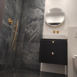 martom-lux - Nowoczesna łazienka z szarymi płytkami imitującymi kamień, złotą armaturą i czarną szafką pod umywalką z okrągłym lustrem z podświetleniem.