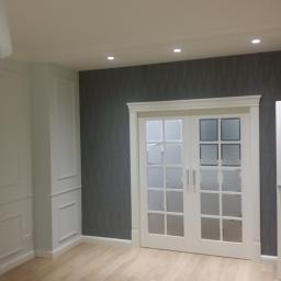 martom-lux - Eleganckie wnętrze z jasną drewnianą podłogą, szarą ścianą z delikatnym wzorem i białymi, przeszklonymi drzwiami przesuwnymi oraz dekoracyjną sztukaterią na ścianach.