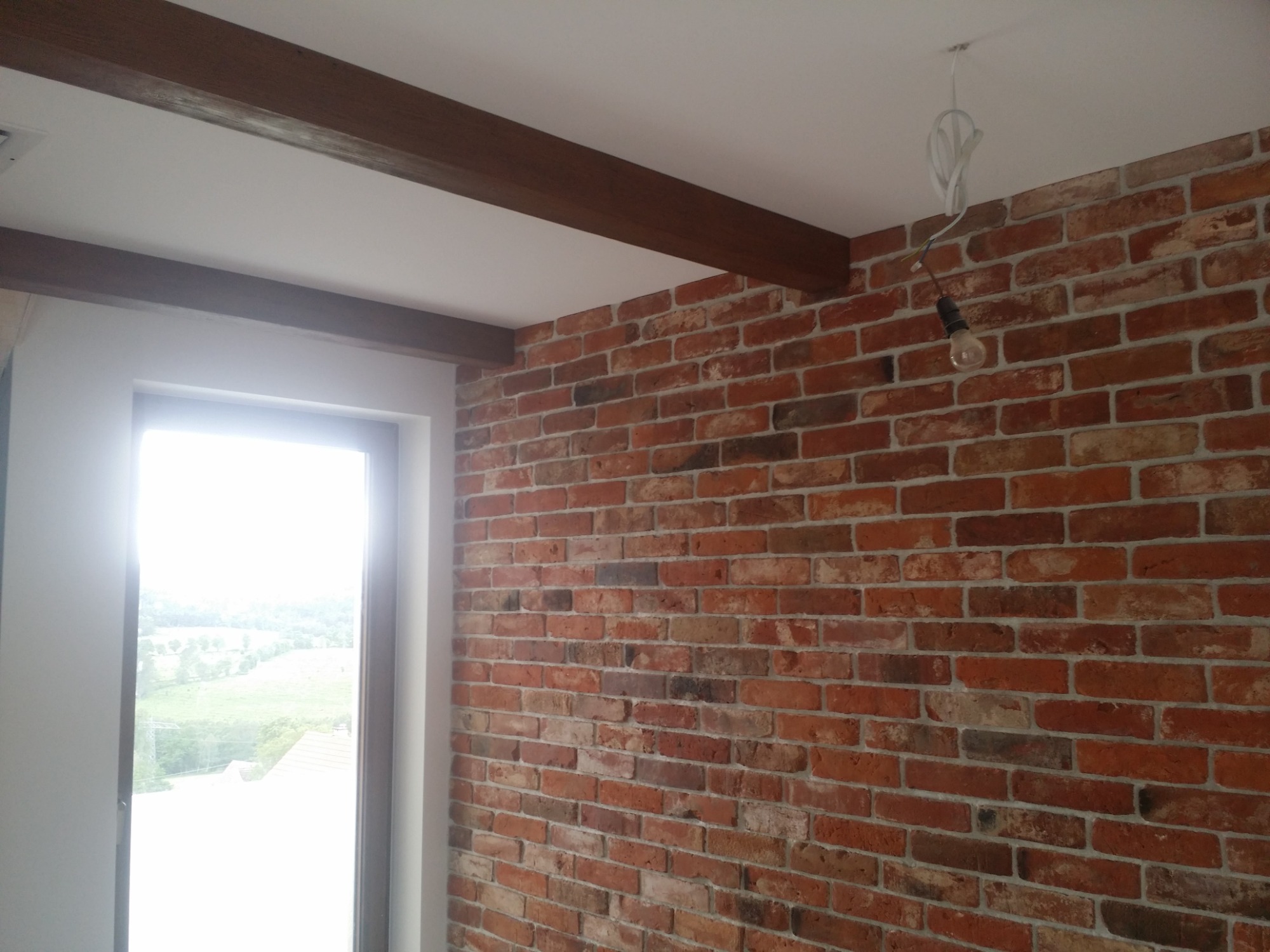 Ściana z czerwonej cegły z dekoracyjnymi belkami stropowymi i wiszącą żarówką bez klosza, widok na zewnątrz przez okno.