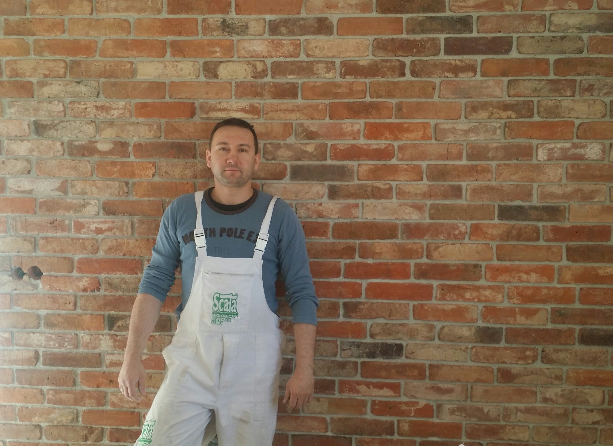 Mężczyzna w białym kombinezonie malarskim z logo firmy stoi na tle ściany z cegły klinkierowej w odcieniach czerwieni i brązu. Widoczne gniazdka elektryczne w ścianie.