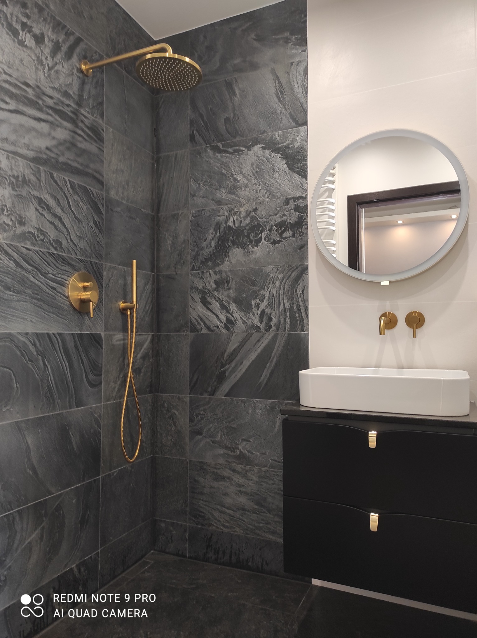 Nowoczesna łazienka z szarymi płytkami imitującymi kamień, złotą armaturą, okrągłym lustrem z podświetleniem LED i czarną szafką pod umywalką.