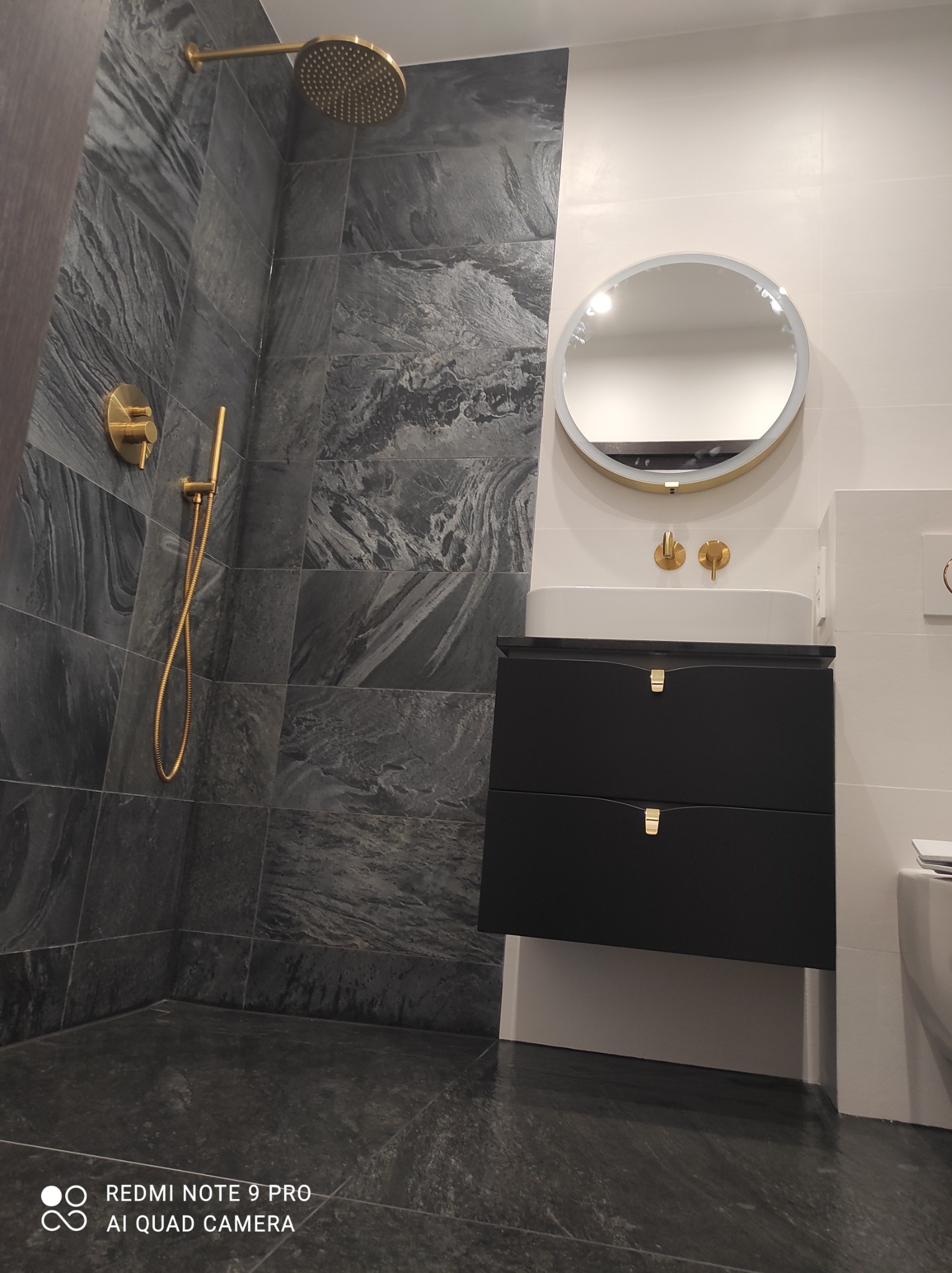 Nowoczesna łazienka z szarymi płytkami imitującymi kamień, złotą armaturą i czarną szafką pod umywalką z okrągłym lustrem z podświetleniem.