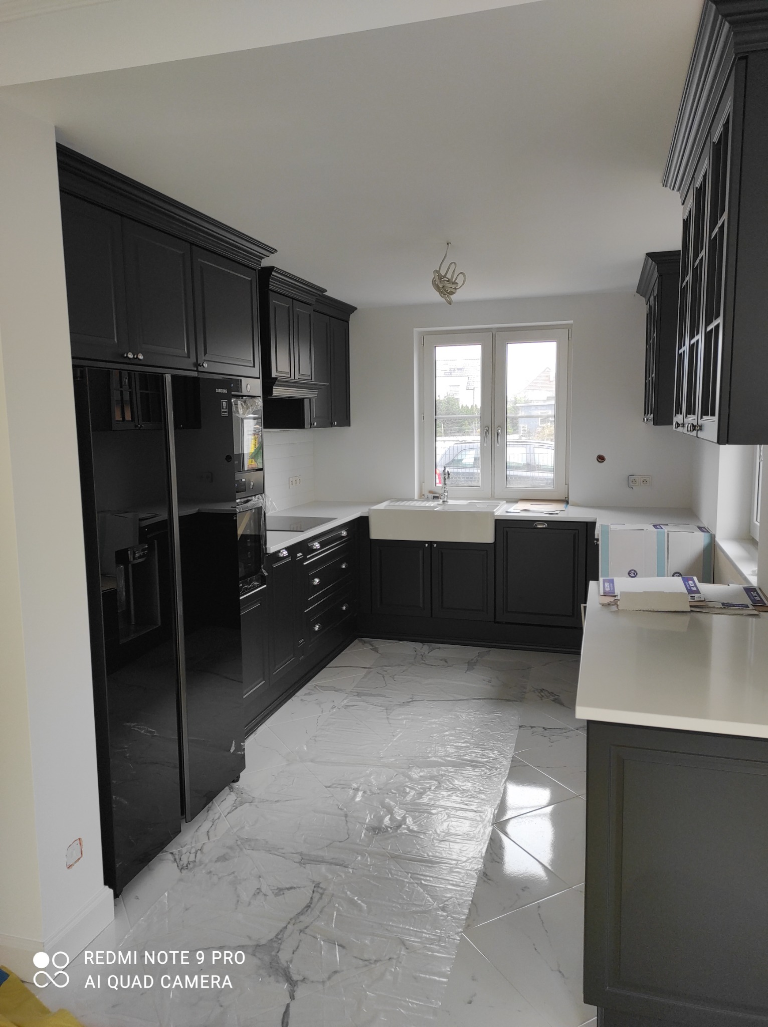 Nowoczesna kuchnia w odcieniach szarości i bieli, z czarnymi szafkami z frezowanymi frontami, białym zlewem farmerskim i marmurową podłogą zabezpieczoną folią ochronną.