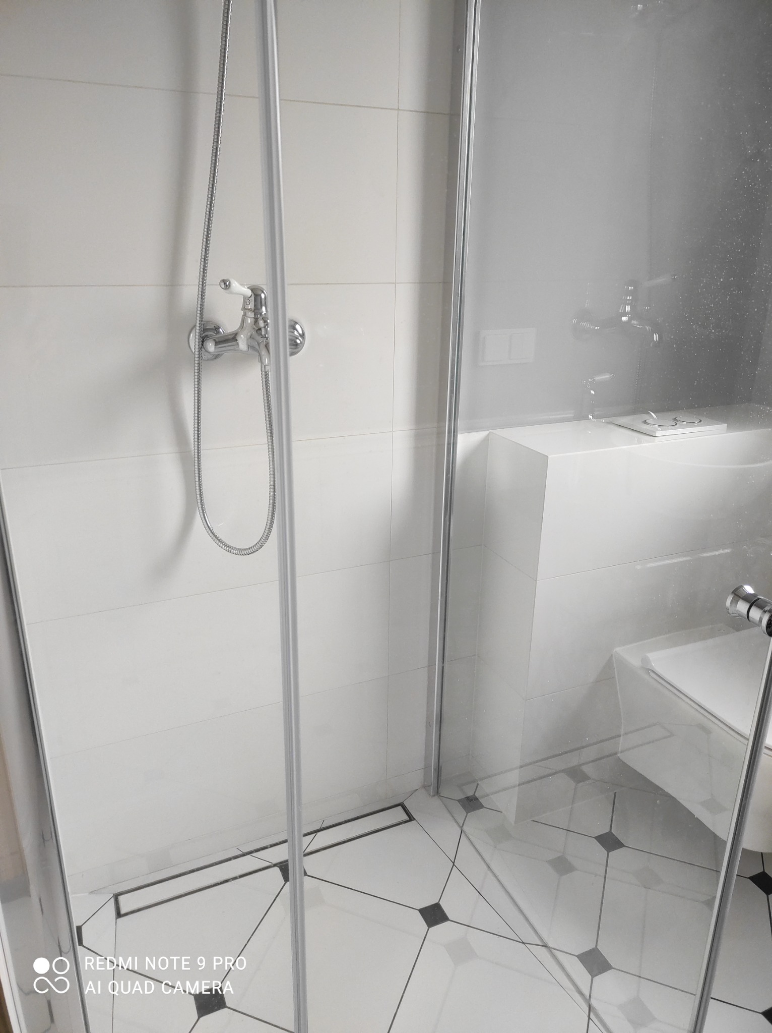 Nowoczesna kabina prysznicowa z chromowaną armaturą i odpływem liniowym, białe płytki ścienne i podłogowe z czarnymi akcentami, minimalistyczna toaleta w tle.