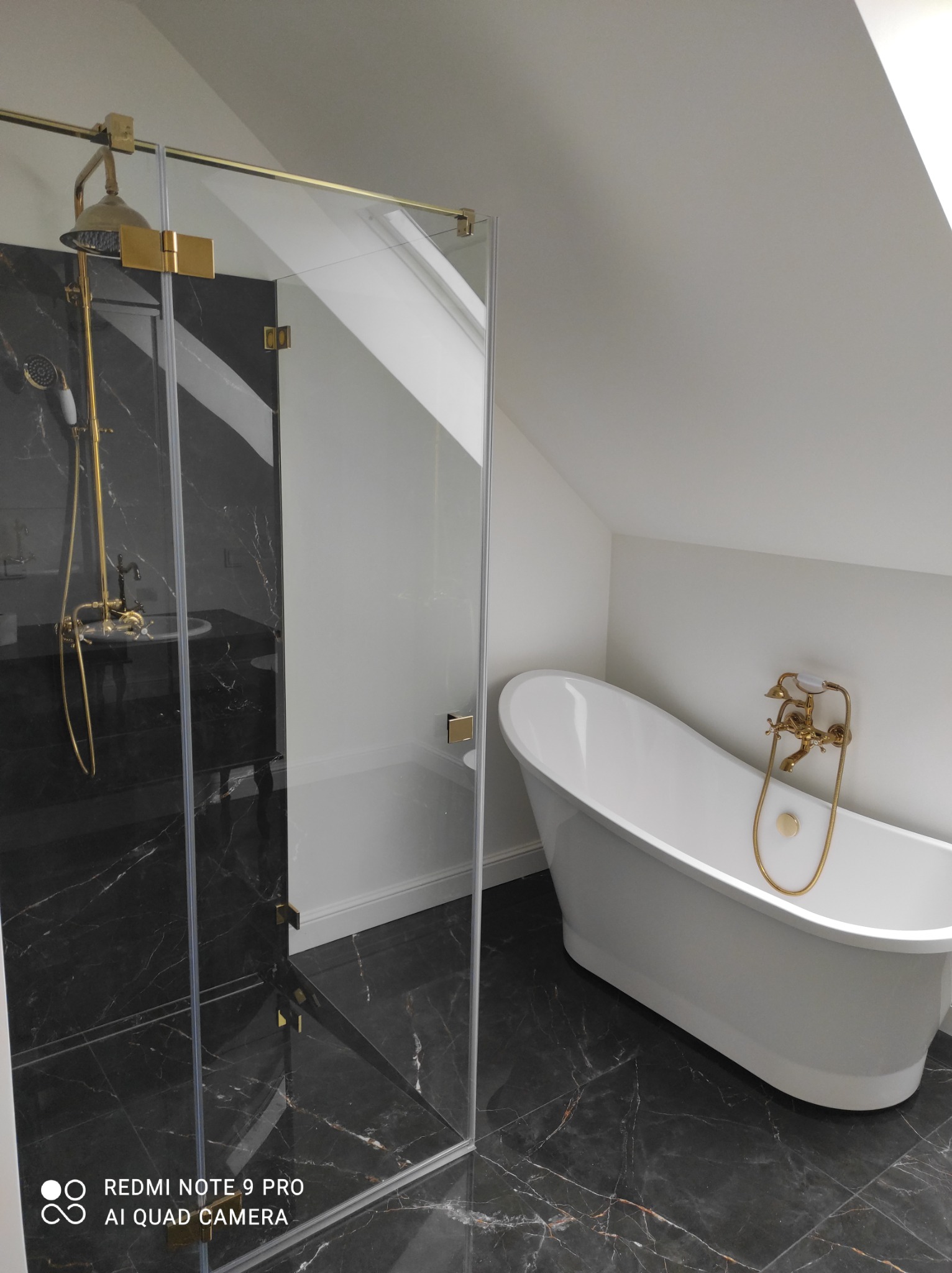 Nowoczesna łazienka na poddaszu z czarną marmurową podłogą, białą wolnostojącą wanną i kabiną prysznicową ze złotymi detalami.