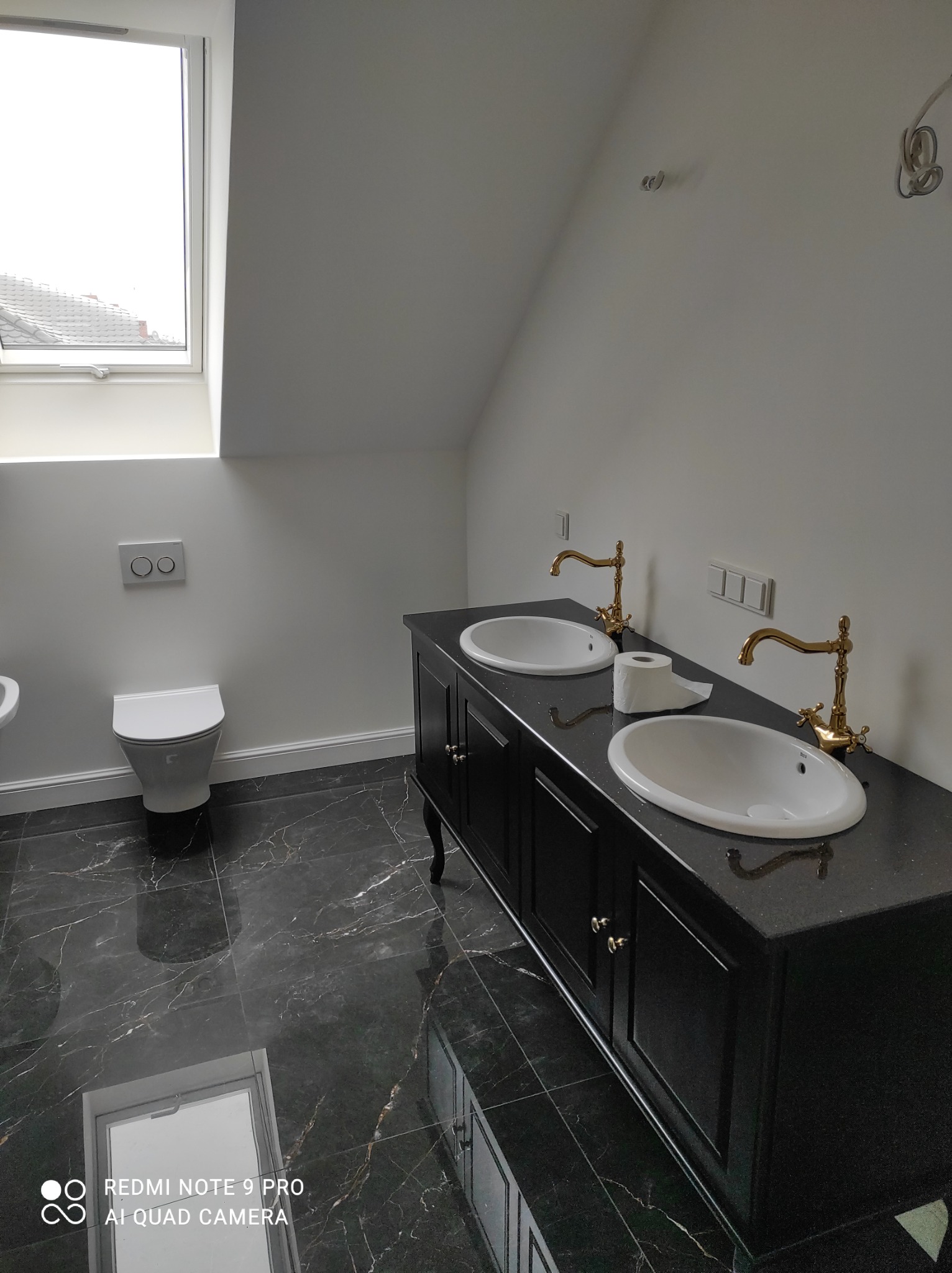 Wykończona łazienka na poddaszu z czarną marmurową podłogą, toaletą z podtynkową spłuczką i szafką pod umywalki z dwiema białymi umywalkami i złotymi bateriami w stylu retro.