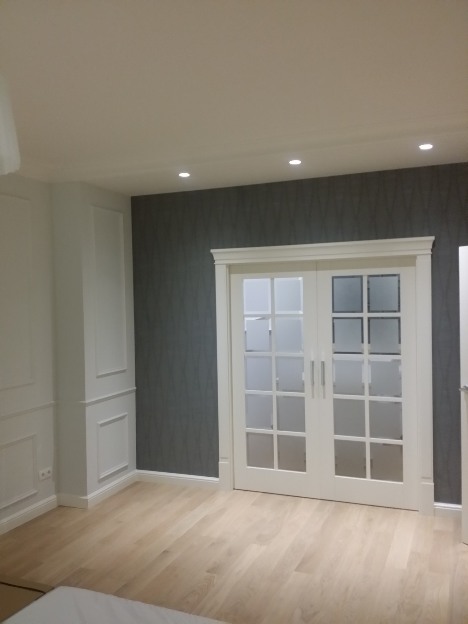 Eleganckie wnętrze z jasną drewnianą podłogą, szarą ścianą z delikatnym wzorem i białymi, przeszklonymi drzwiami przesuwnymi oraz dekoracyjną sztukaterią na ścianach.