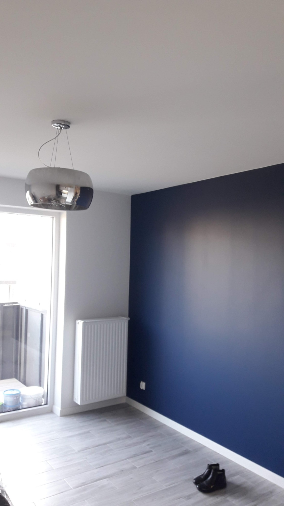 Pomalowane na granatowo wnętrze pokoju z grzejnikiem, lampą wiszącą i podłogą imitującą drewno, z parą czarnych butów na podłodze.