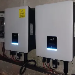 Dwa białe falowniki fotowoltaiczne marki FOX, zamontowane na ścianie z betonową fakturą, obok skrzynki elektrycznej. Widoczne przewody podłączeniowe i listwy montażowe.