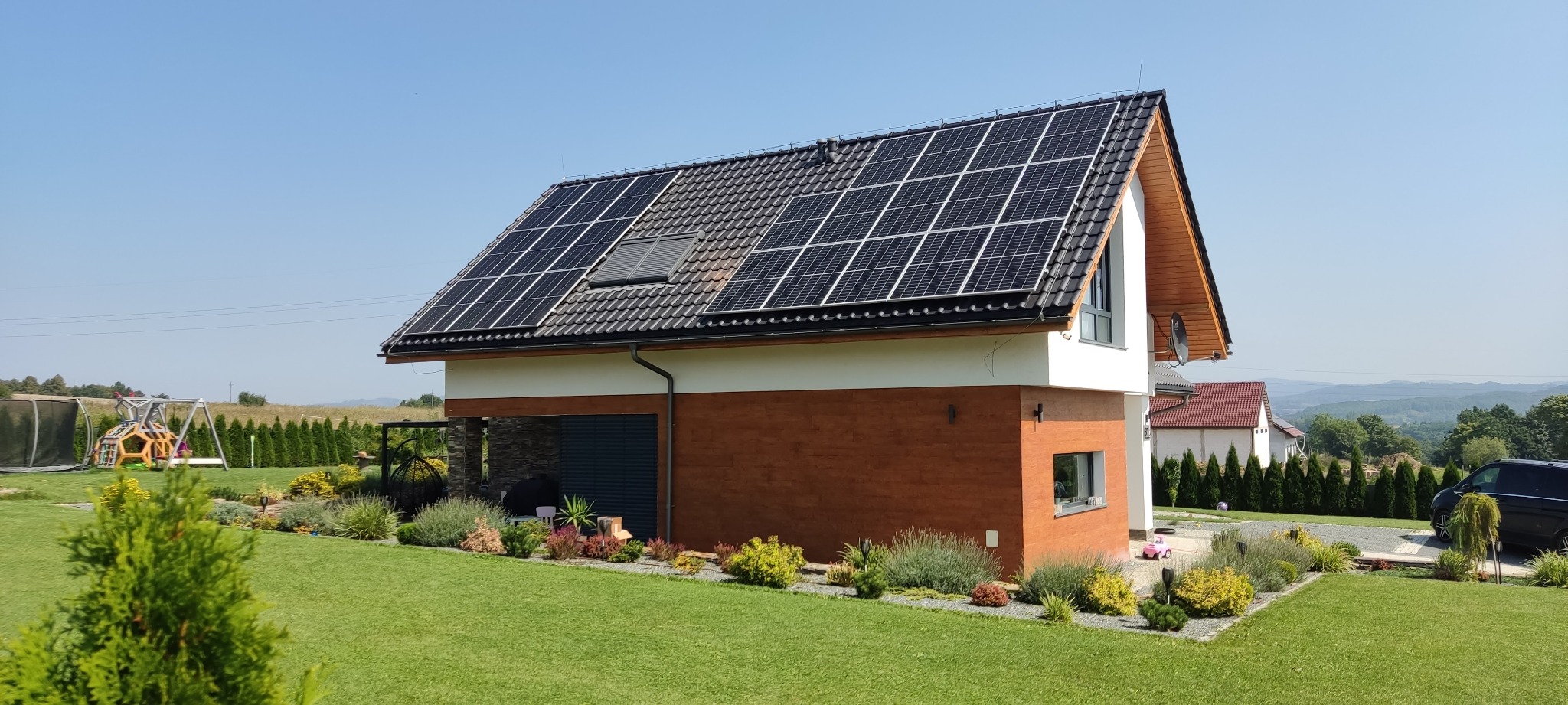 Dom jednorodzinny z panelami słonecznymi na dachu, otoczony zadbanym ogrodem z widokiem na pagórkowaty krajobraz.