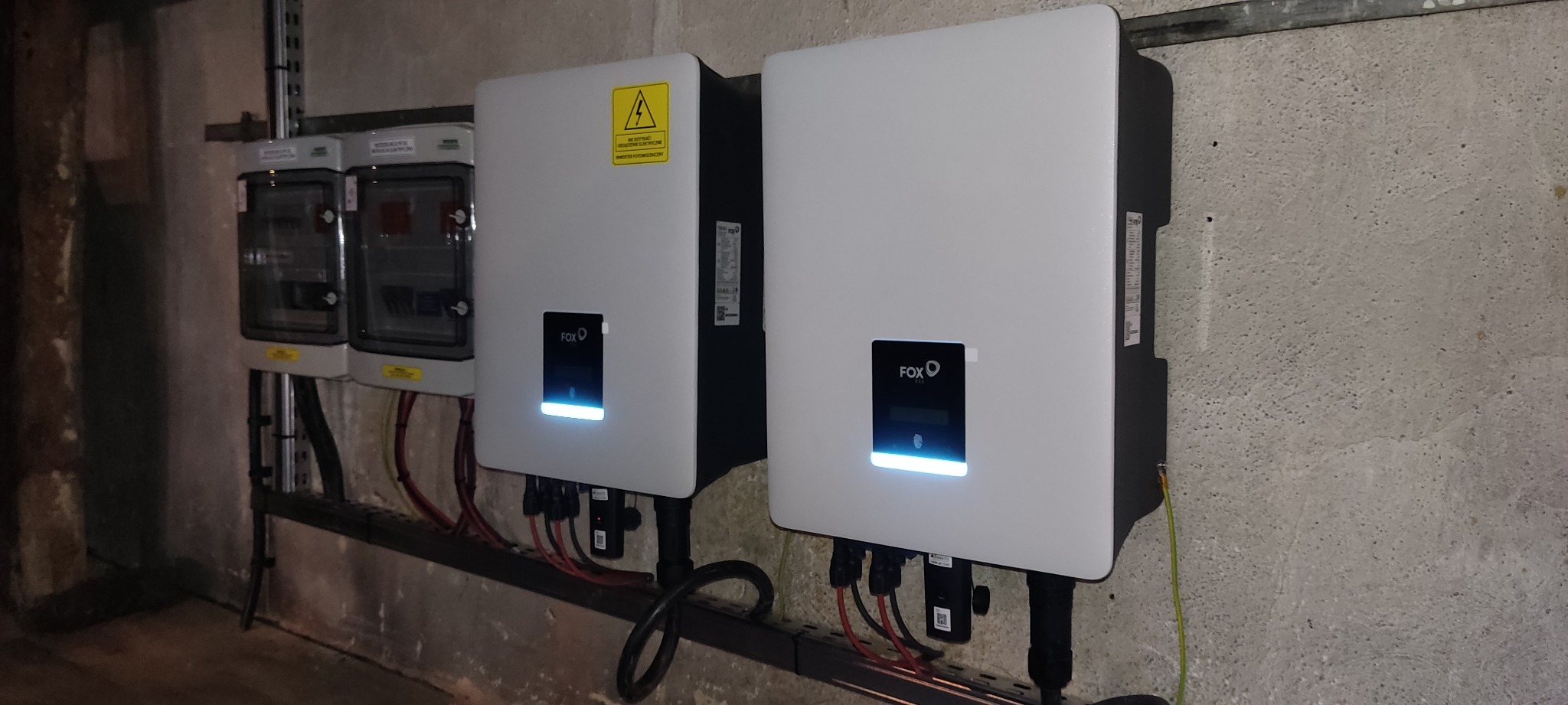 Dwa białe falowniki fotowoltaiczne marki FOX, zamontowane na ścianie z betonową fakturą, obok skrzynki elektrycznej. Widoczne przewody podłączeniowe i listwy montażowe.