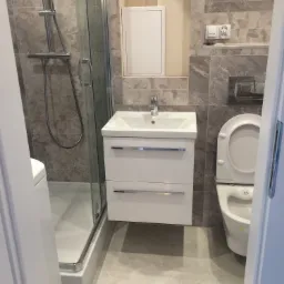 Nowoczesna łazienka z kabiną prysznicową, umywalką z szafką, toaletą i szarą płytką imitującą marmur na ścianach i podłodze, widziana zza uchylonych drzwi.