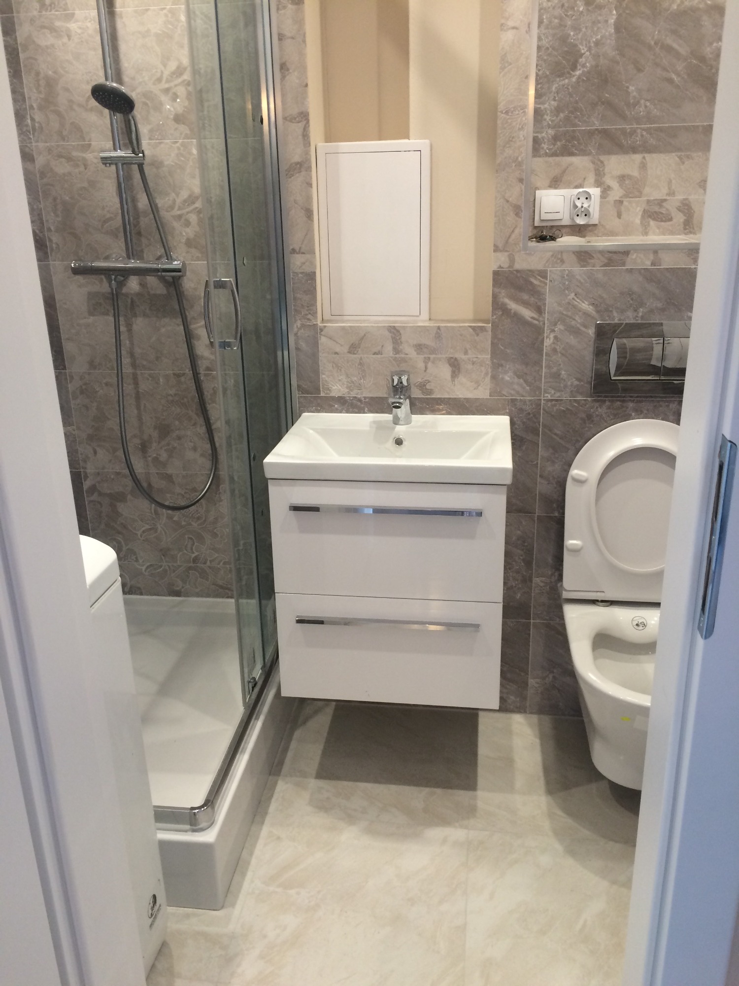 Nowoczesna łazienka z kabiną prysznicową, umywalką z szafką, toaletą i szarą płytką imitującą marmur na ścianach i podłodze, widziana zza uchylonych drzwi.