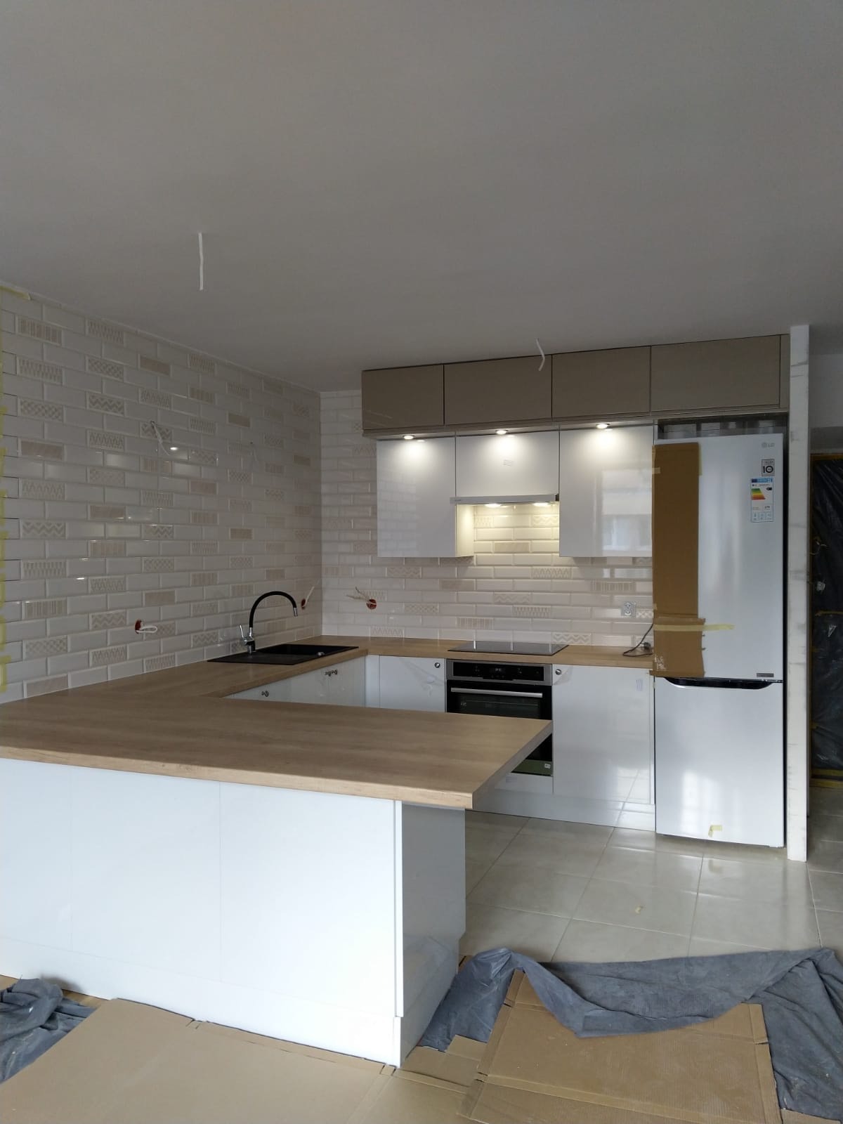 Wnętrze kuchni w trakcie remontu: białe meble z drewnianym blatem, płytki ścienne w stylu cegiełki, widoczne niedokończone prace i zabezpieczenia na podłodze.