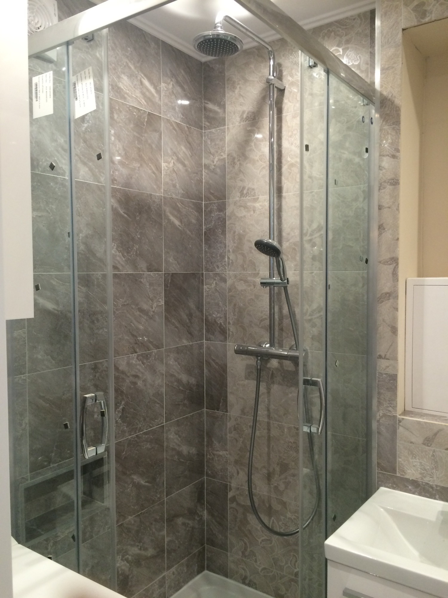 Kabina prysznicowa z przesuwnymi drzwiami ze szkła hartowanego, wykończona szarymi płytkami imitującymi marmur, z chromowaną armaturą i deszczownicą.