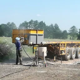 Pracownik w kombinezonie ochronnym czyści konstrukcję naczepy ciężarowej za pomocą urządzenia do obróbki strumieniowo-ściernej; widoczny pył i maszyna do piaskowania.