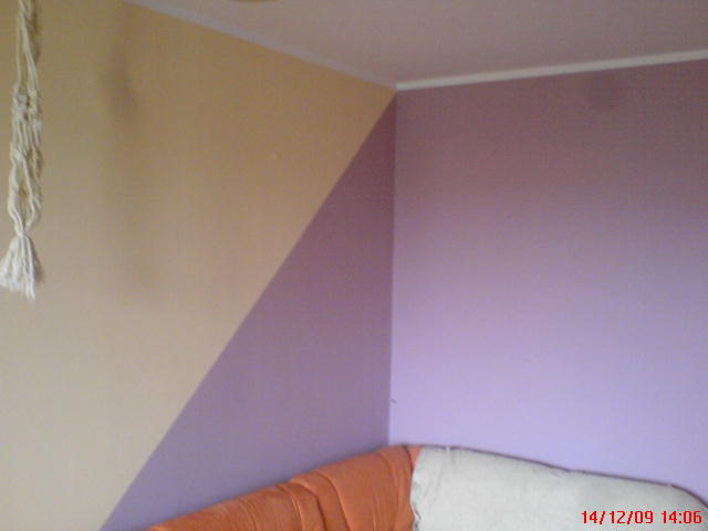 Pomieszczenie z pomalowanymi ścianami w dwóch kolorach: beżowym i fioletowym, rozdzielonych ukośną linią. Na ścianie wisi makrama. Fragment pomarańczowej sofy widoczny w dolnej części kadru.