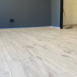 MichBud - Jasne panele podłogowe ułożone w pokoju z pomalowanymi na szaro ścianami, zabezpieczonymi niebieską taśmą malarską przy podłodze. W tle widoczny fragment pomieszczenia z płytkami.