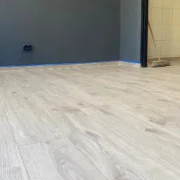 Jasne panele podłogowe ułożone w pokoju z pomalowanymi na szaro ścianami, zabezpieczonymi niebieską taśmą malarską przy podłodze. W tle widoczny fragment pomieszczenia z płytkami.