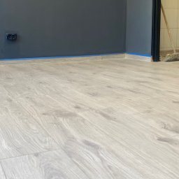 MichBud - Jasne panele podłogowe ułożone w rogu pokoju z szarą ścianą, zabezpieczoną niebieską taśmą malarską. Widoczny fragment listwy przypodłogowej i gniazdko elektryczne.