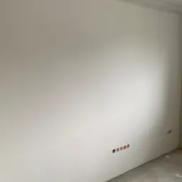 Ściana po szpachlowaniu i malowaniu na biało z widocznymi gniazdkami elektrycznymi i fragmentem ściany w stanie surowym w rogu pomieszczenia.