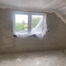 MichBud - Pokój w trakcie szpachlowania ścian, z oknem zabezpieczonym folią i widocznymi śladami prac na podłodze. Ściany częściowo pokryte szarą masą, sufit w trakcie wykańczania.