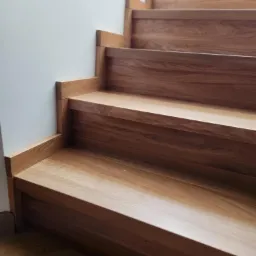 Detal drewnianych schodów dębowych z bliska, widoczne usłojenie i subtelne oświetlenie. Minimalistyczny design, jasna ściana w tle.