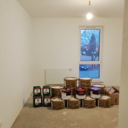 MichBud - Puszki z farbą i gruntem Atlas w pustym, jasnym pokoju z oknem. Przygotowania do prac wykończeniowych. Widok na zewnątrz przez okno.