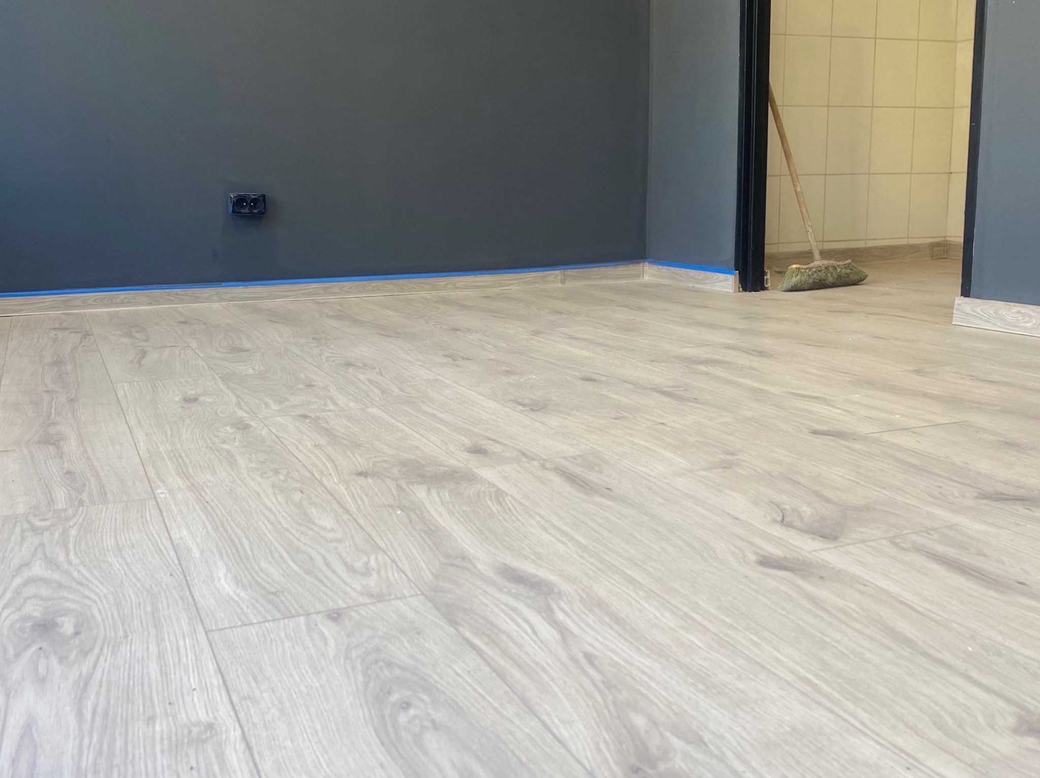 Jasne panele podłogowe ułożone w pokoju z pomalowanymi na szaro ścianami, zabezpieczonymi niebieską taśmą malarską przy podłodze. W tle widoczny fragment pomieszczenia z płytkami.