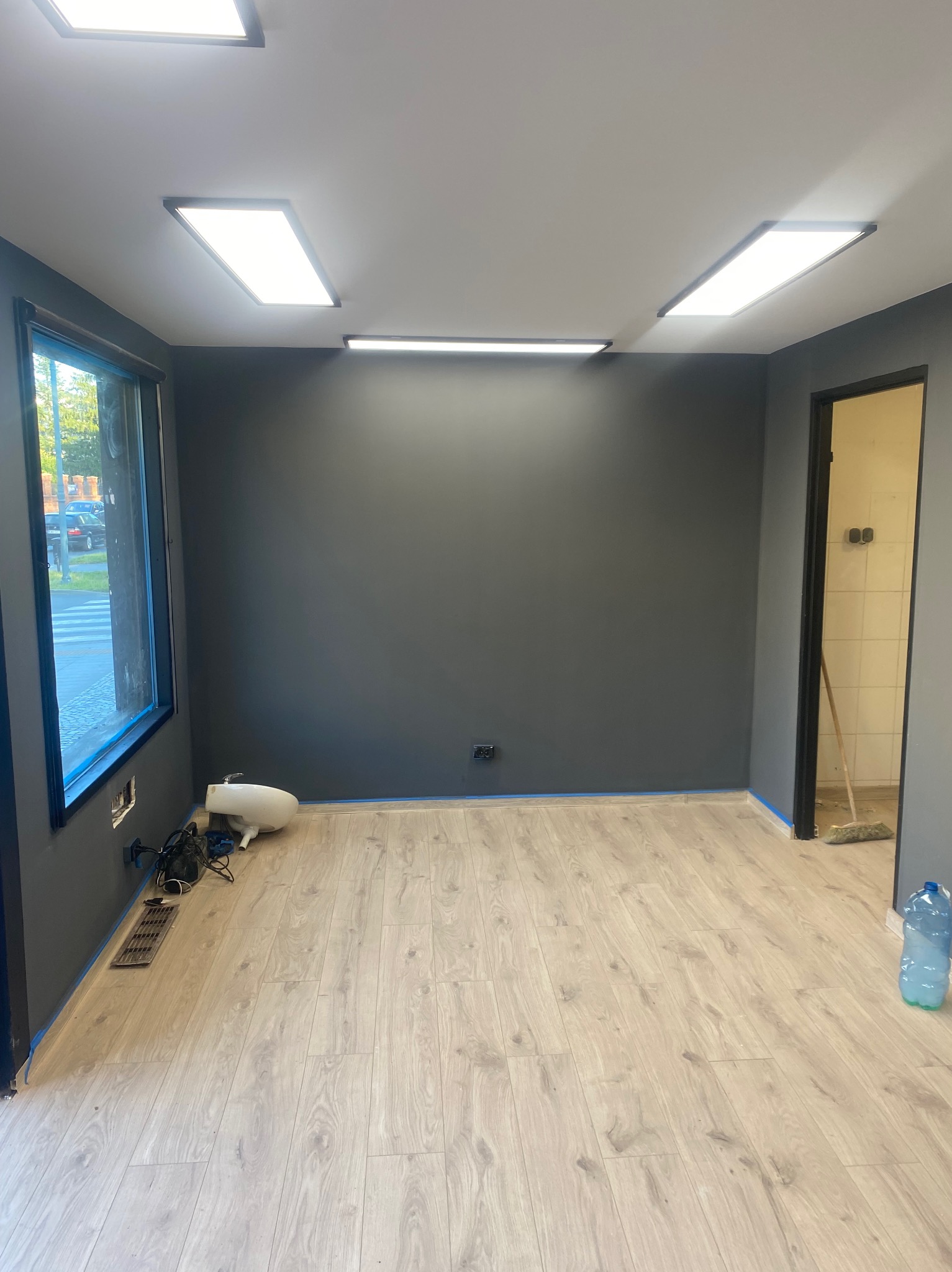 Wnętrze po malowaniu na szaro, z jasnymi panelami podłogowymi i niebieską taśmą malarską przy podłodze. Widoczne okno i otwarte drzwi do pomieszczenia z jasnymi płytkami.