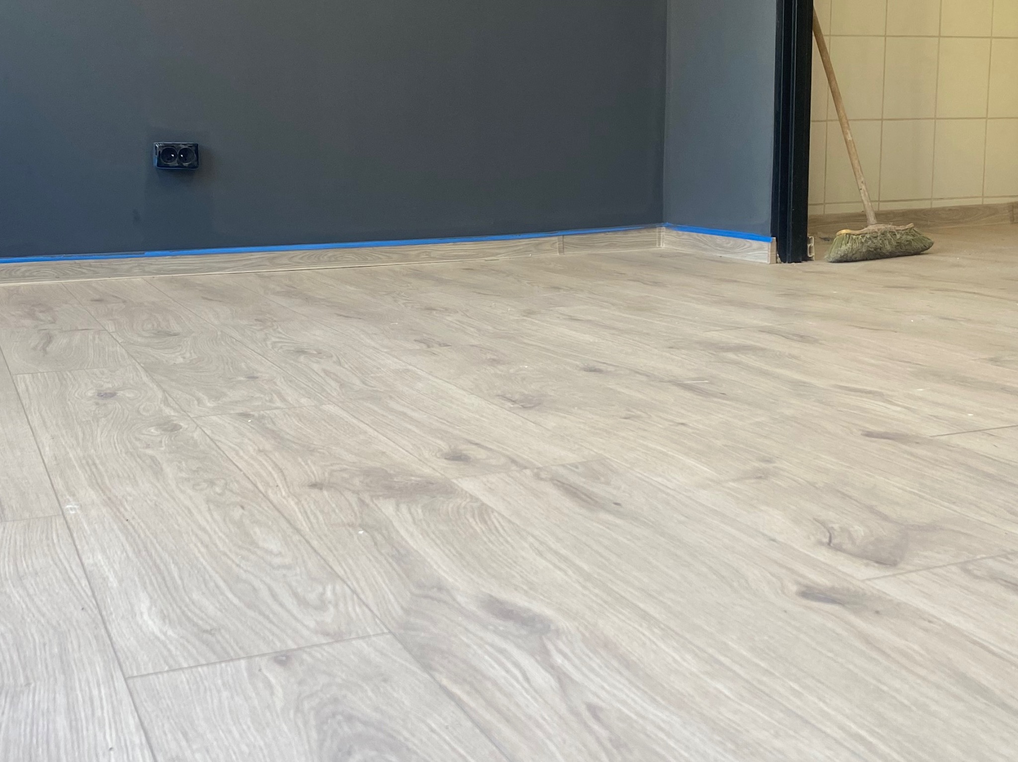Jasne panele podłogowe ułożone w rogu pokoju z szarą ścianą, zabezpieczoną niebieską taśmą malarską. Widoczny fragment listwy przypodłogowej i gniazdko elektryczne.