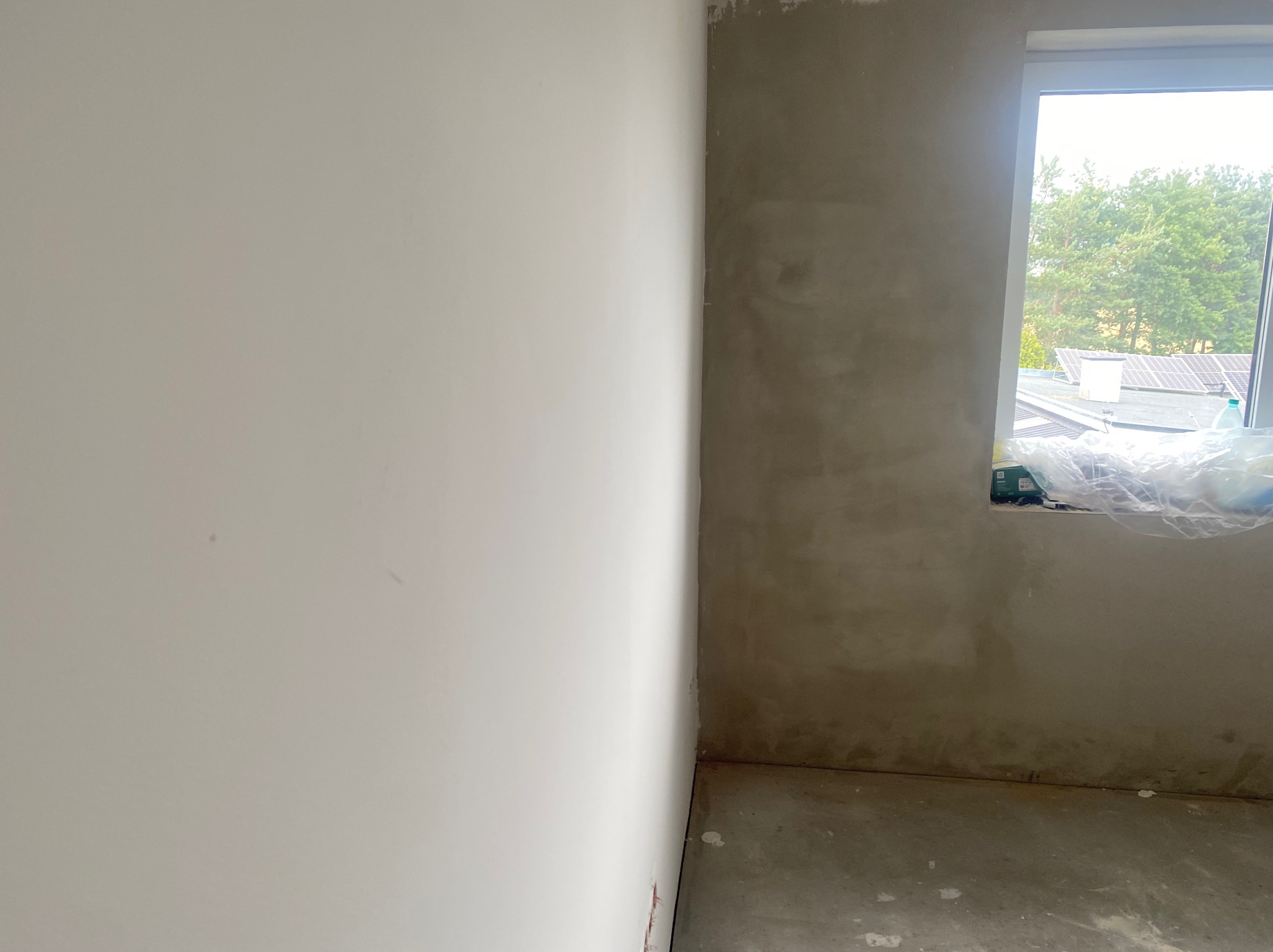 Fragment wnętrza: biała ściana po lewej, surowa ściana po prawej z oknem. Widoczne przygotowania do dalszych prac wykończeniowych.