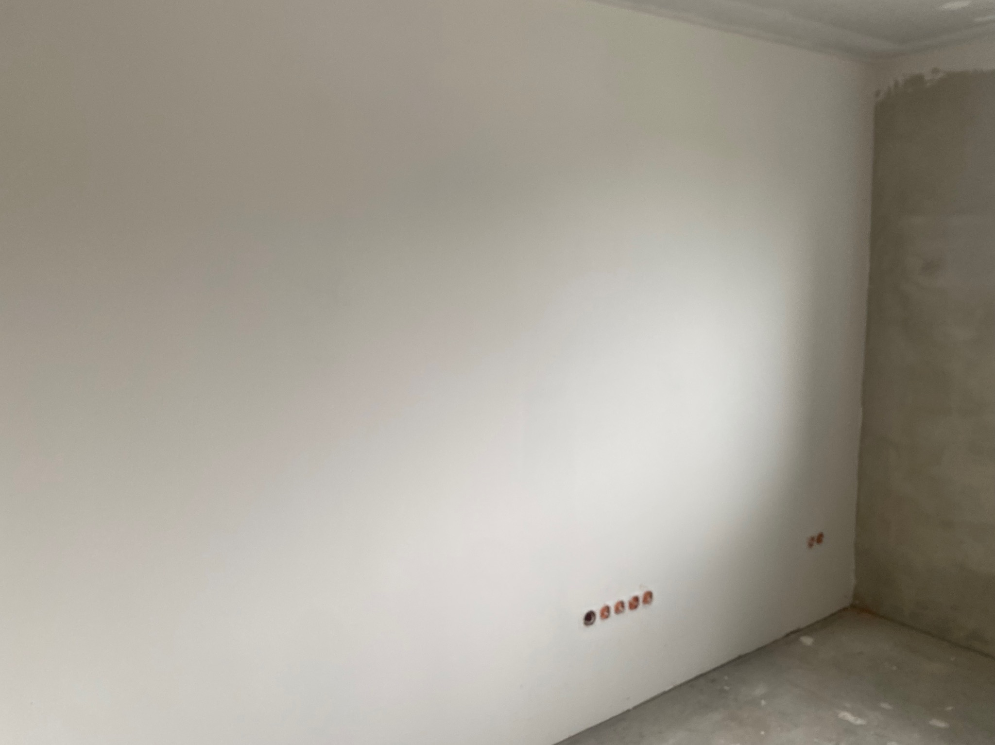 Ściana po szpachlowaniu i malowaniu na biało z widocznymi gniazdkami elektrycznymi i fragmentem ściany w stanie surowym w rogu pomieszczenia.