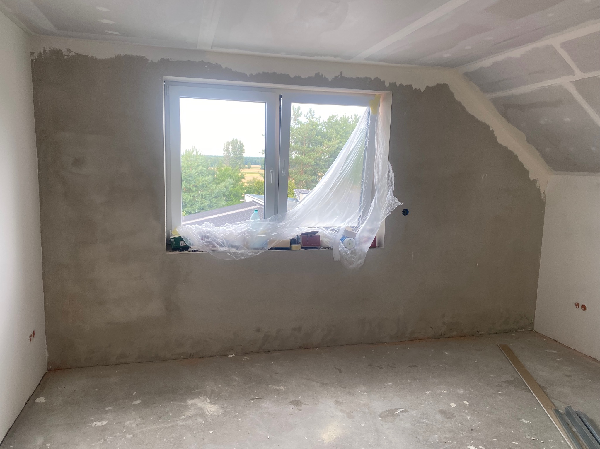 Pokój w trakcie szpachlowania ścian, z oknem zabezpieczonym folią i widocznymi śladami prac na podłodze. Ściany częściowo pokryte szarą masą, sufit w trakcie wykańczania.