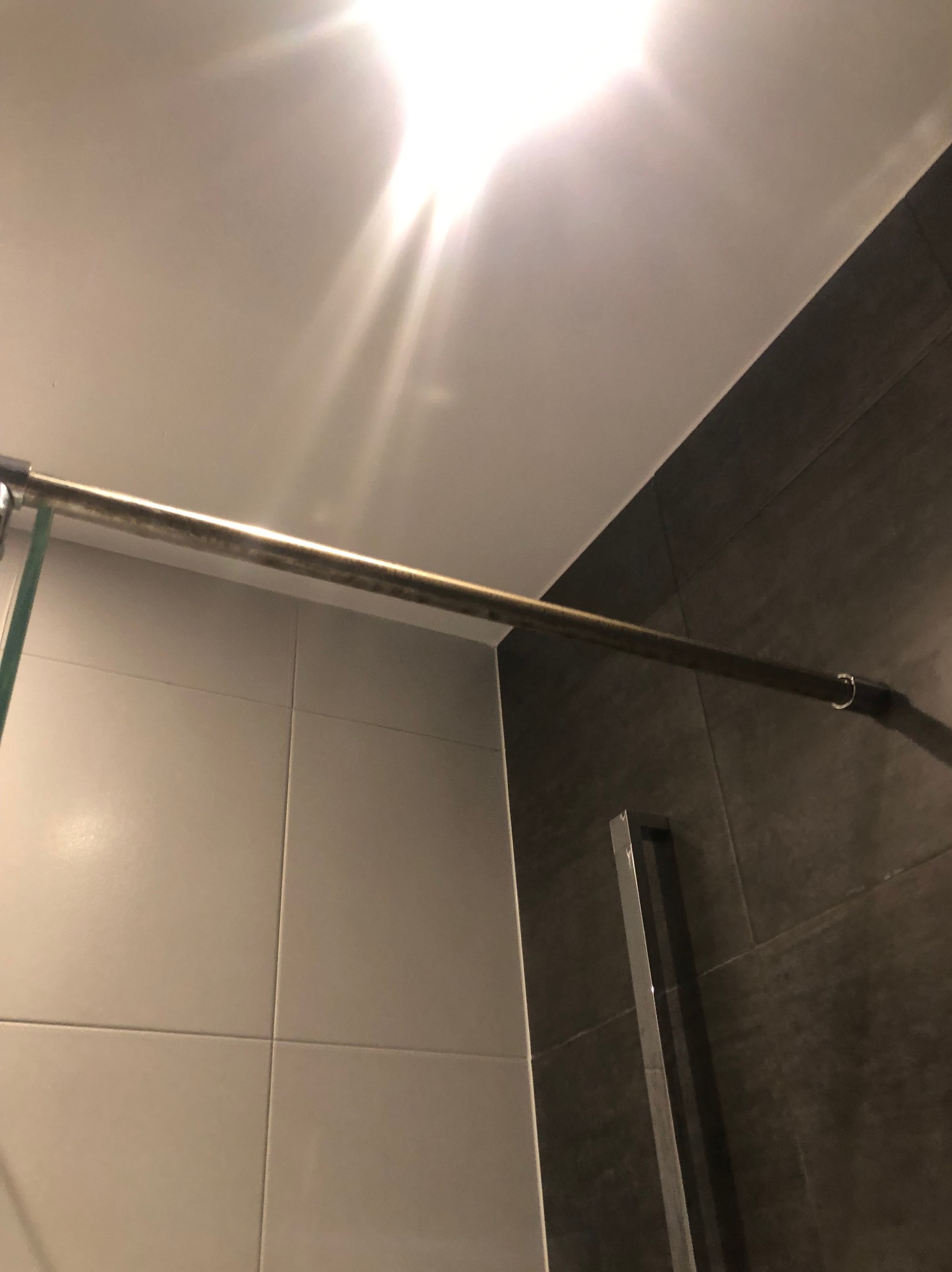 Fragment łazienki z kabiną prysznicową: białe i grafitowe płytki, metalowy reling. Ujęcie z dołu, widoczny sufit i mocne oświetlenie.